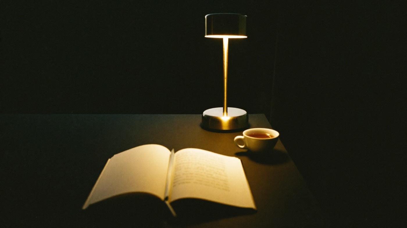 An open book and a cup of tea in the warm glow of a single brass lamp on a dark desk
